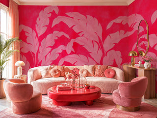 Bold pink banana leaf wall mural on red background in a cozy living room with curved blush sofa and gold mirror