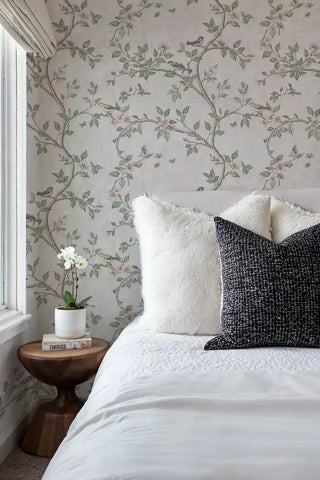 Botanical wallpaper with delicate birds and pink flowers on branches, shown in serene bedroom with white bedding and round nightstand