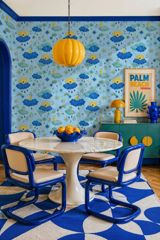 Celestial wallpaper with evil-eye suns and blue clouds in a dining room with tulip table and yellow pendant