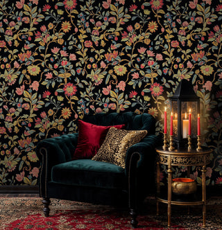 Colorful floral tapestry wallpaper on black background, styled in a moody living room with green velvet armchair and red candles