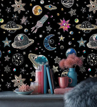 Cosmic galaxy wallpaper with jeweled planets and sparkling stars on black, shown with decorative vase and books