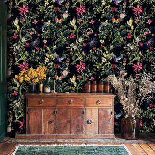 Dark botanical wallpaper featuring hummingbirds and pink flowers, in rustic room with wooden sideboard and dried flowers