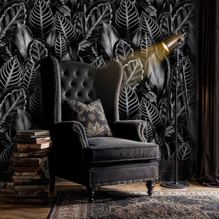 Dark botanical wallpaper with lush white-edged leaves, behind a tufted wingback armchair and stacked books