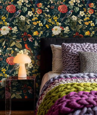 Dark botanical wallpaper with songbirds and flowers, shown in cozy bedroom with mushroom lamp and knit throws
