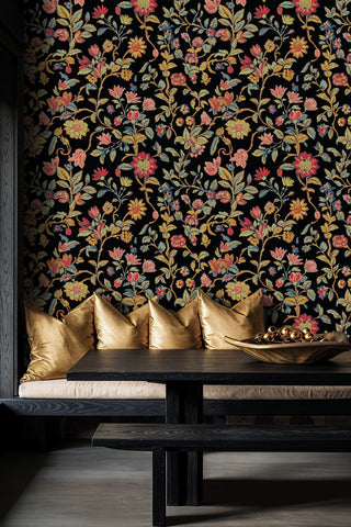 Dark embroidered floral wallpaper with red and gold botanicals on black, shown in a modern dining nook with gold cushions and dark bench