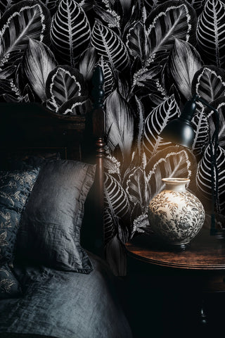 Dark tropical leaf wallpaper with white-veined foliage on black, in a moody bedroom with ceramic vase