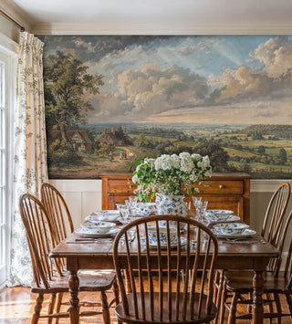 Detailed view of a pastoral landscape wall mural with cottages, hedgerows and big sky in green and blue, classic painted style, shown by a farmhouse dining table set with blue-and-white china