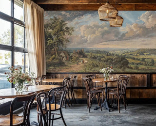 Detailed view of a pastoral landscape wall mural with cottages, hedgerows and open sky in green and blue, classic painted style, shown in a cafe with rattan pendants and bistro chairs