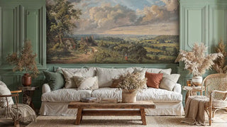 Detailed view of a pastoral landscape wall mural with cottages, rolling hills and dramatic clouds in green and blue, classic painted style, shown with a linen sofa and rustic coffee table