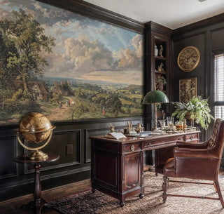 Detailed view of a pastoral landscape wall mural with cottages, valley fields and sunlit clouds in green and blue, classic painted style, shown in a study with a brass globe and leather desk chair