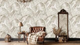 Elegant tropical wallpaper with oversized leaves in neutral tones, displayed in traditional living room with leather armchair