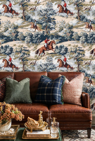 Equestrian scenic wallpaper with horses, riders and wooded hills, beige and blue, behind a brown leather sofa