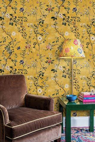 Golden botanical wallpaper with wild blooms and dark foliage, styled in a living room with brown velvet armchair and books