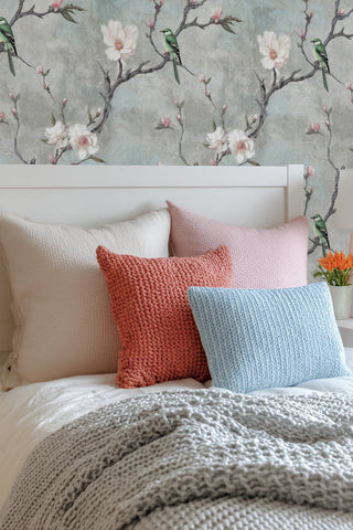 Grey magnolia wallpaper with green birds shown in cozy bedroom with colorful knit pillows