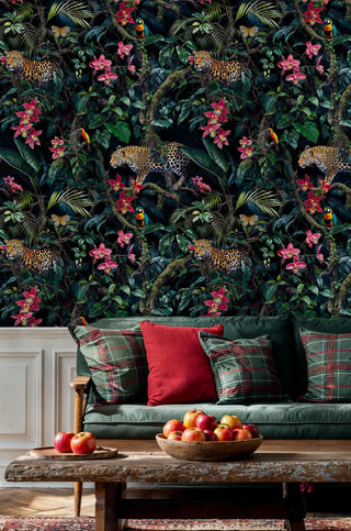 Jungle wallpaper with leopards, parrots and pink orchids on dark, shown in a living room with plaid cushions and rustic table apples