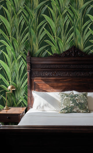 Lush green leaf wallpaper with golden accents behind an ornate carved wood bed and brass lamp