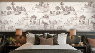 Pastoral toile wall mural in warm brown on ivory, shown above a tufted headboard in a cozy bedroom with green accent pillows