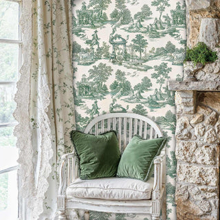 Scenic toile wallpaper in sage green and cream, shown in cottage bedroom with white chair and stone wall