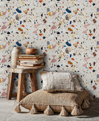 Terrazzo wallpaper with colorful stone fragments in pink, navy and orange, shown in cozy reading nook with woven stool