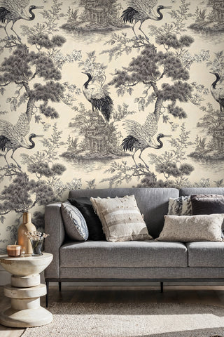 Toile crane wallpaper featuring pine branches and pagoda in ink on cream, in a living room with grey sofa
