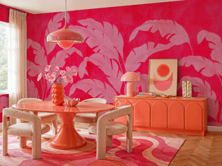 Tropical banana leaf wall mural in red and pink shown in a retro dining room with coral pedestal table and arched sideboard